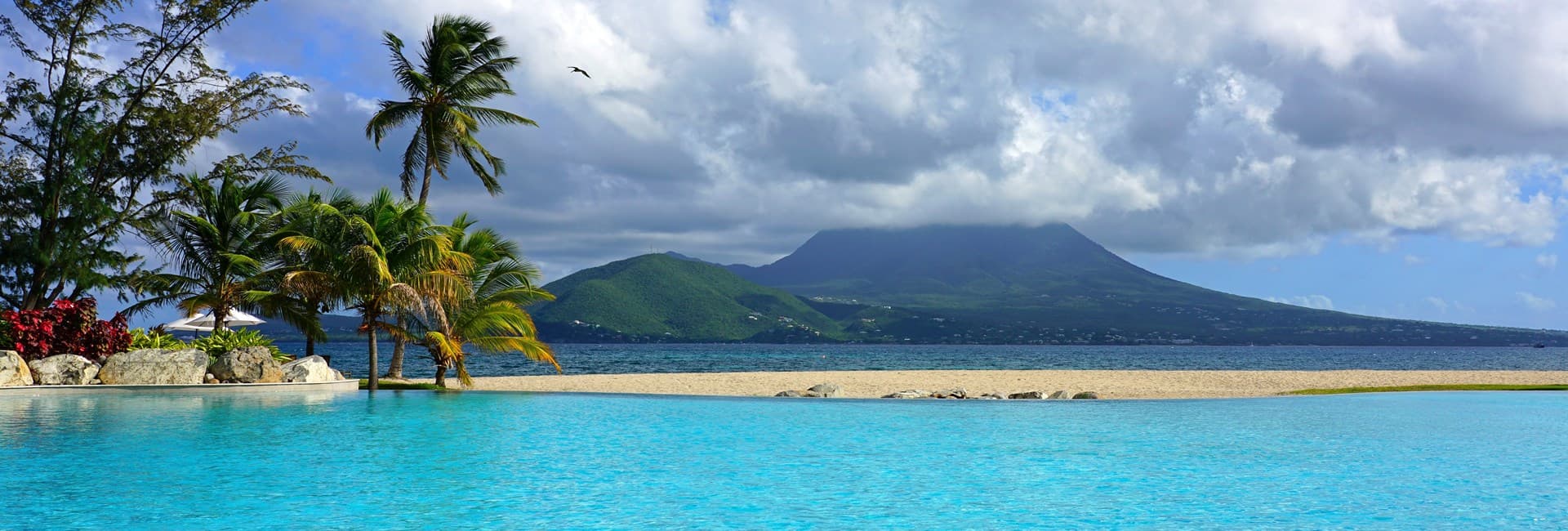A turquoise infinity pool overlooks a sandy beach and palm trees, with a large green mountain rising from the ocean in the distance under a cloudy sky.