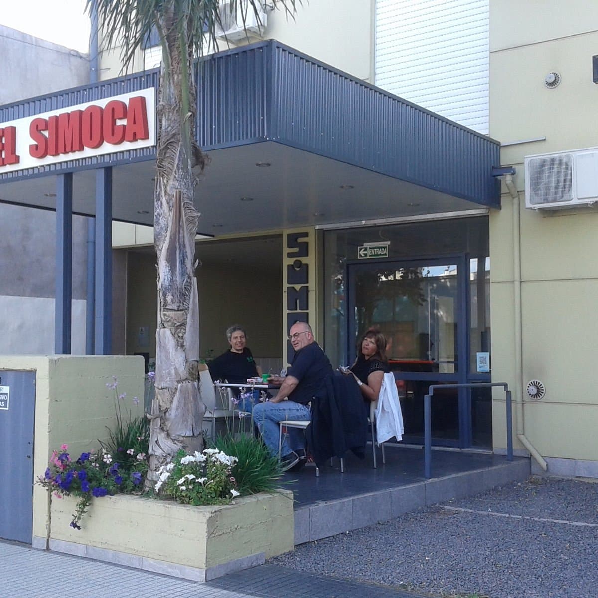 Exterior view of a building with a sign reading 'EL SIMOCA', featuring three people seated at a table under an awning near a palm tree.