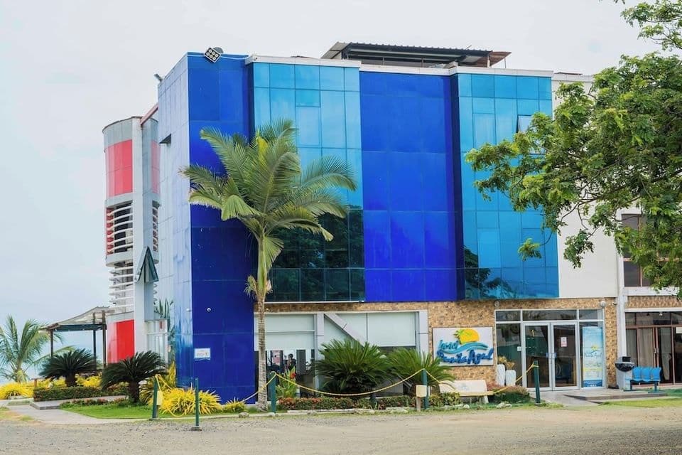 Exterior view of Hotel Punta Azul in Pedernales, Ecuador, with blue glass panels, a red tower, palm trees, and landscaping.