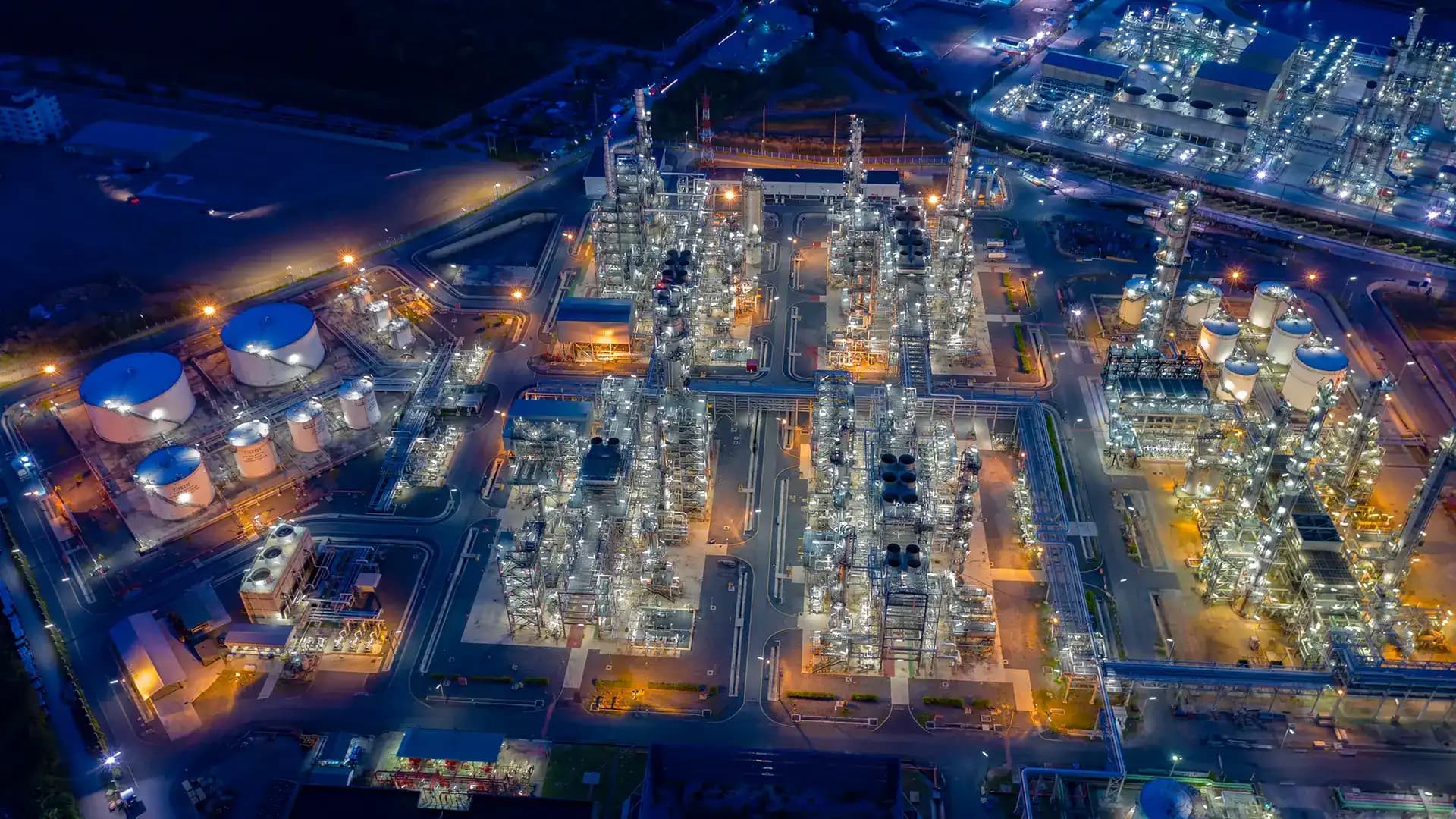 Aerial night view of a sprawling industrial complex featuring illuminated processing towers, storage tanks, and intricate piping networks.