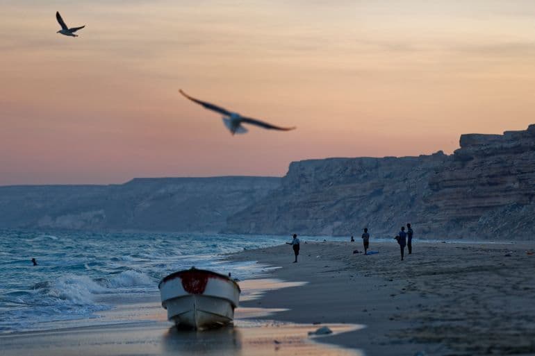 Dusk coastal scene with a small boat on the wet sand, seagulls in flight, and distant figures on the beach.