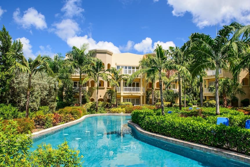 A sunny exterior view of a yellow resort building with balconies, surrounded by palm trees and a winding swimming pool.