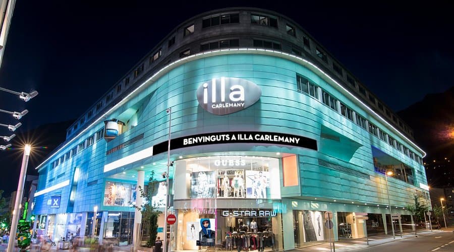 Night view of the Illa Carlemany shopping center in Les Escaldes, featuring illuminated turquoise architecture and storefronts.