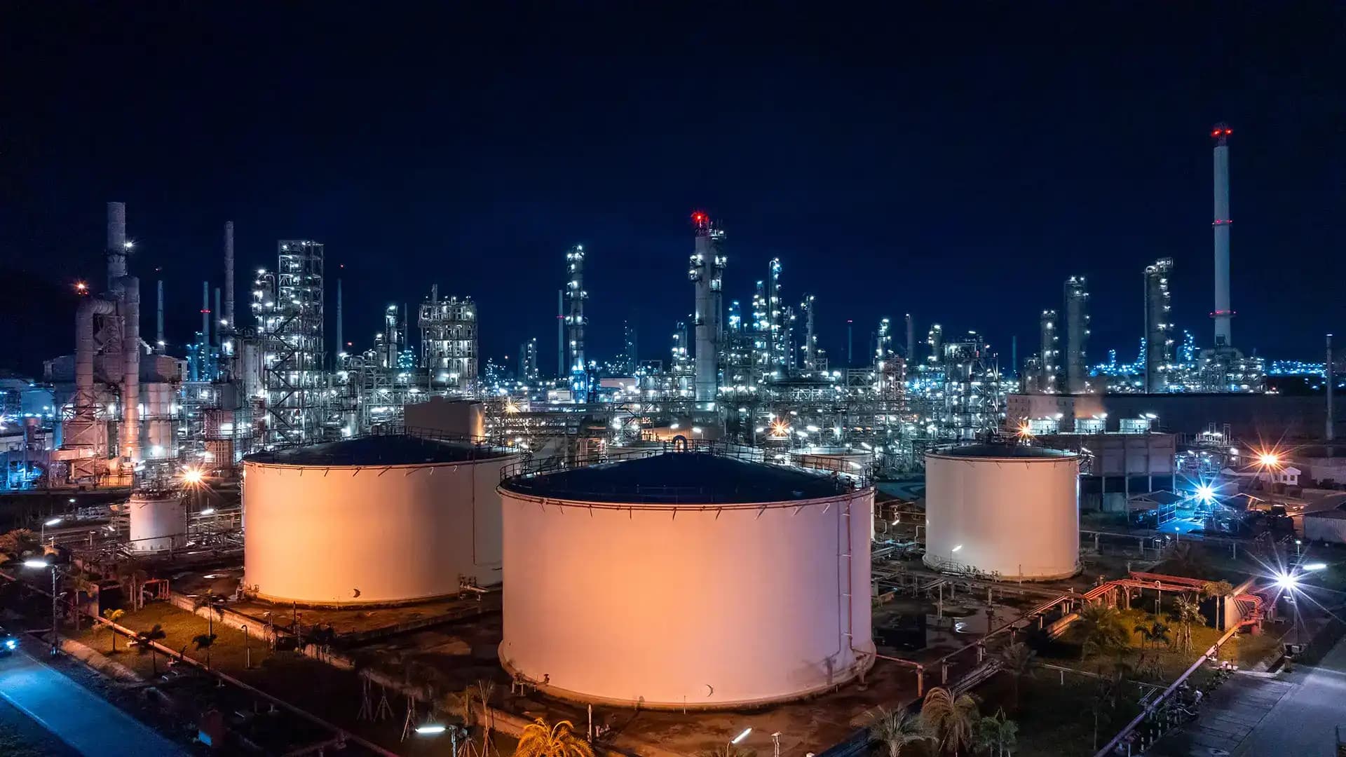 Nighttime aerial view of a sprawling industrial complex featuring large illuminated storage tanks and intricate refinery structures against a dark sky.