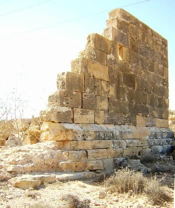 Sunlit exterior of ancient stone ruins in a desert landscape, featuring weathered masonry blocks and a standing wall section.