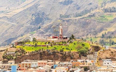 A scenic view of a town in Catarama, Ecuador, featuring a prominent monument on a grassy hill against a backdrop of arid mountains.