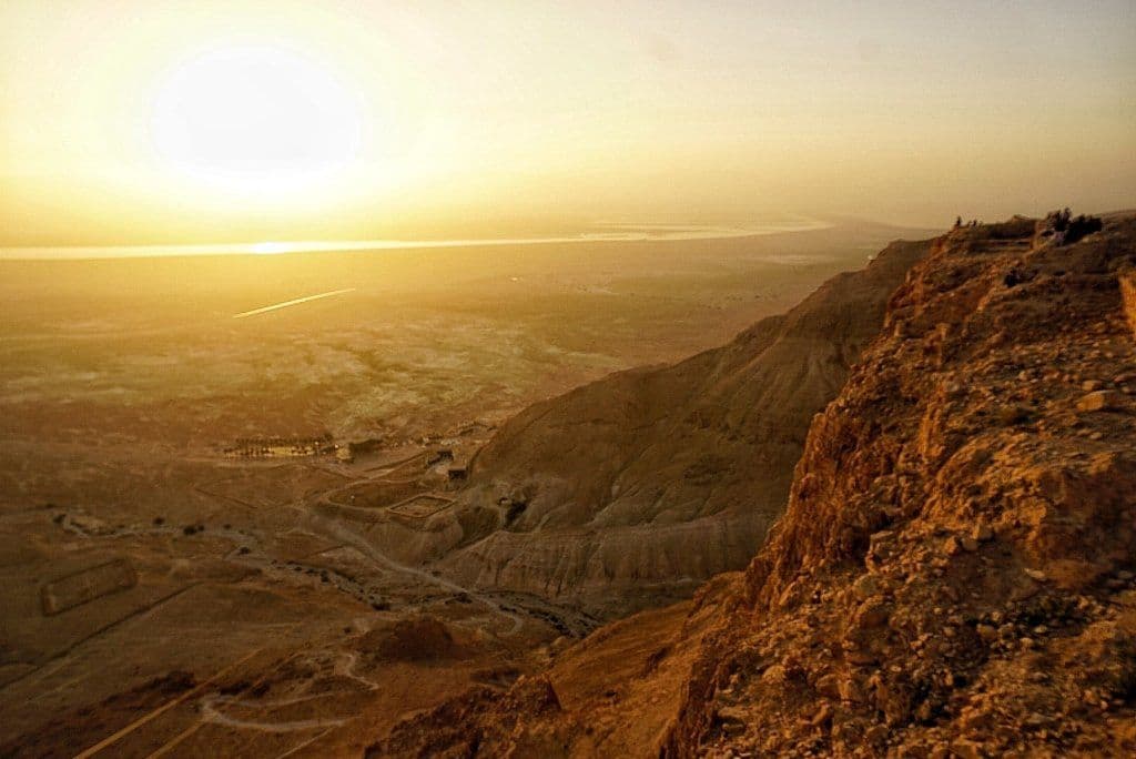 Golden-hour view from a rocky hillside overlooking a desert valley, distant water, and a small settlement under a hazy sky in the central Israel region.
