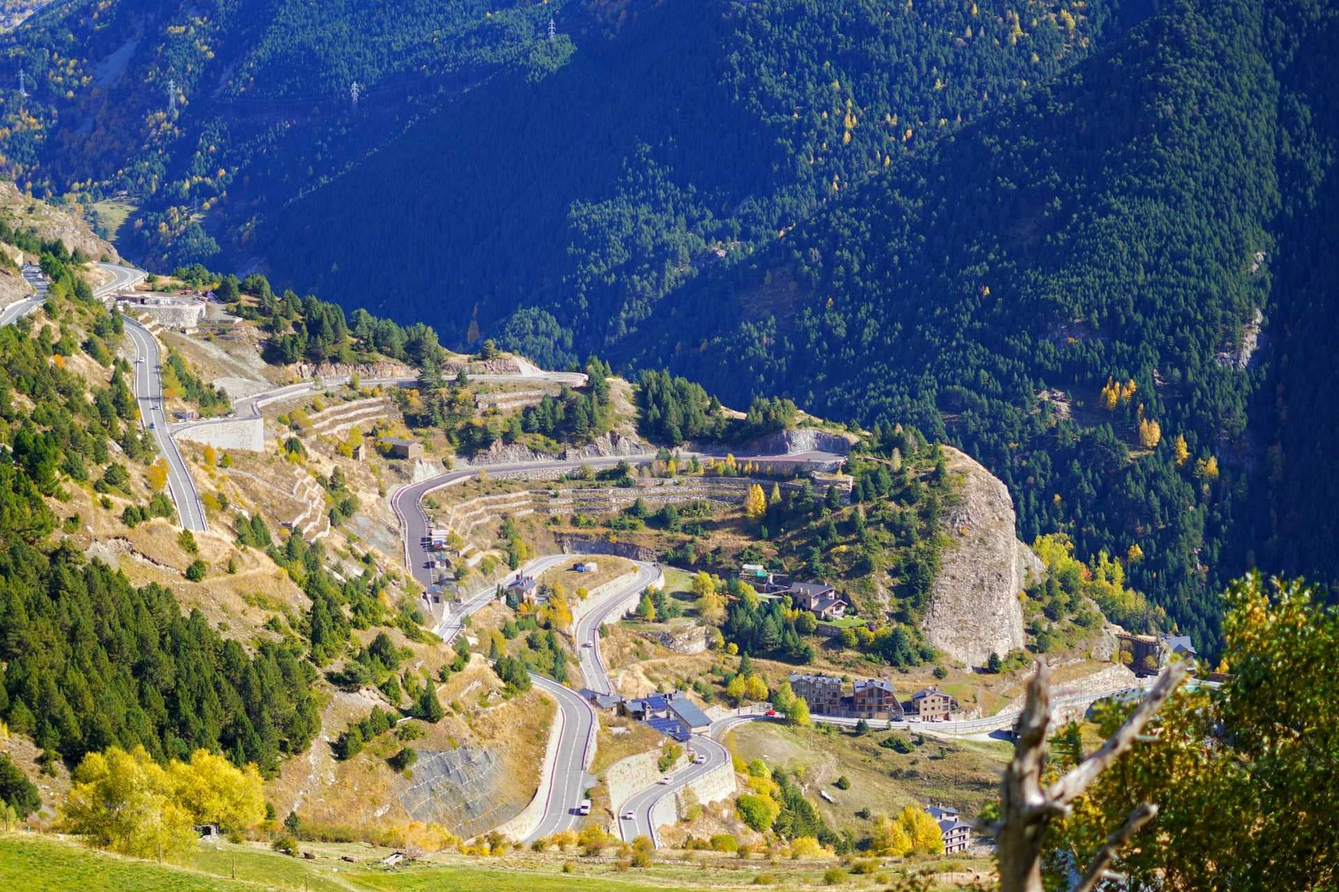 Winding mountain road with hairpin turns descends through forested slopes toward a small village in autumn.