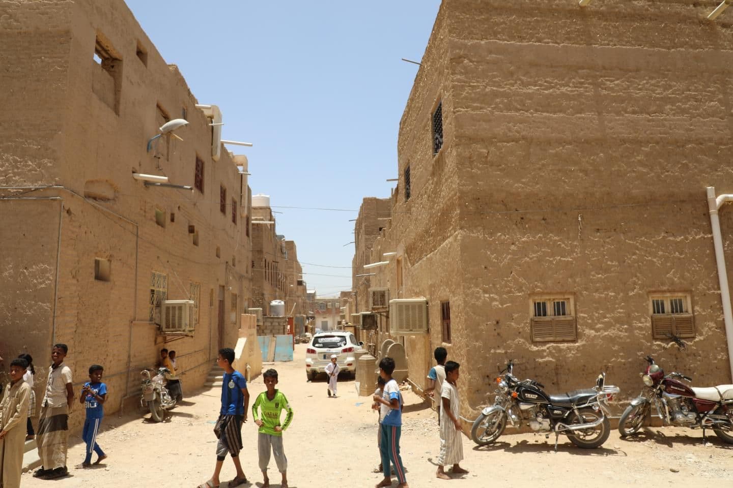 Children and locals stand in a sunlit, narrow street lined with traditional mud-brick buildings and parked motorcycles in the Hadhramaut region.