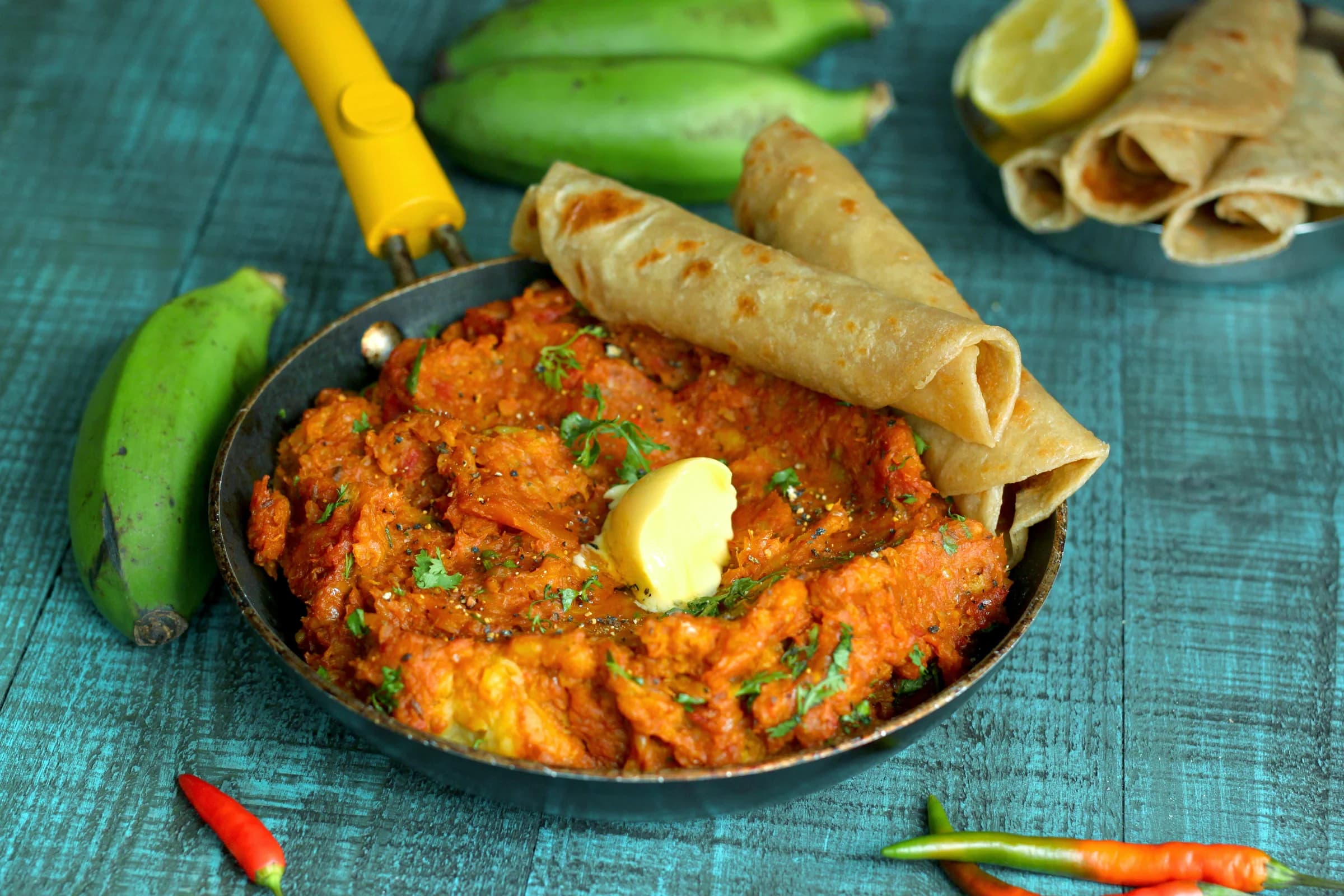 A skillet of vibrant orange Ugandan matooke stew topped with butter and herbs, served with rolled flatbreads on a teal wooden table.