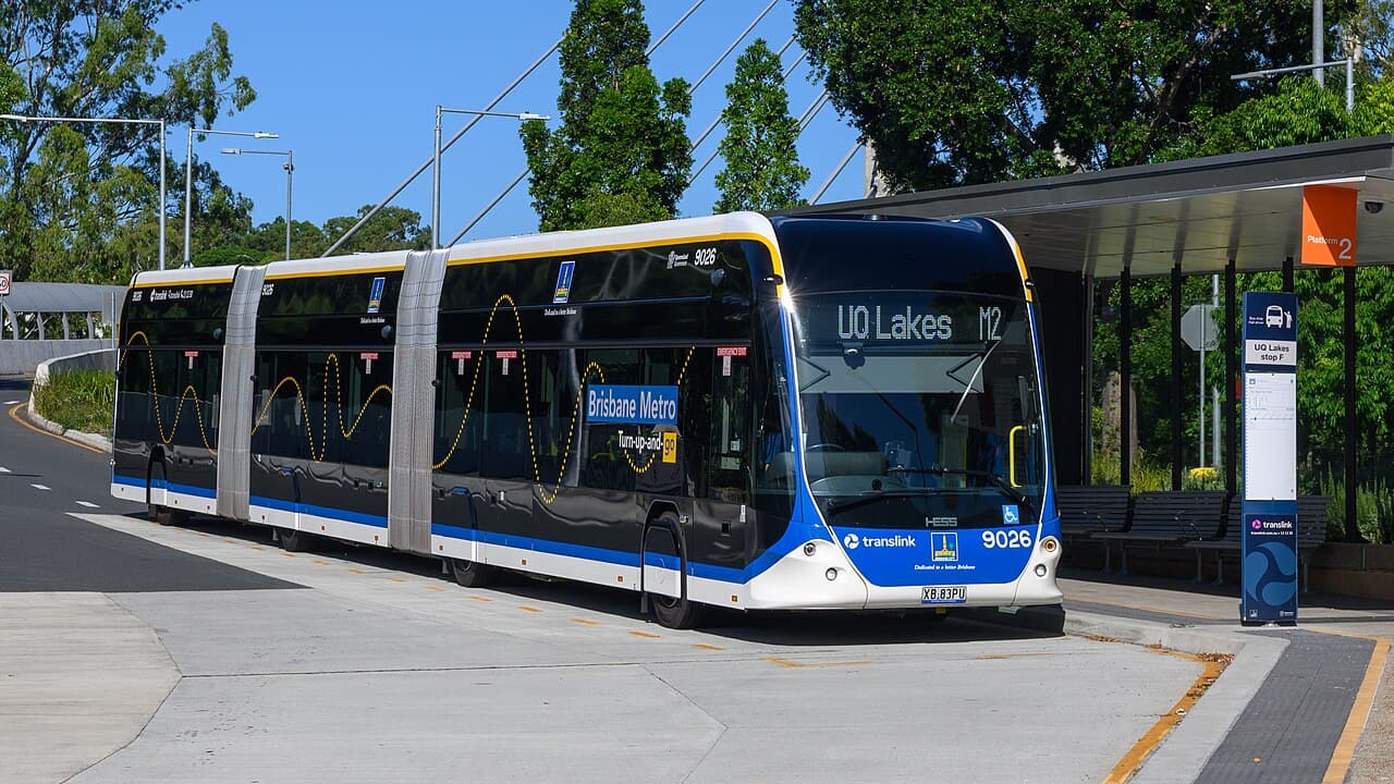 A blue and white Translink articulated bus, route UQ Lakes, is parked at a modern bus station platform in Brisbane under a clear blue sky.