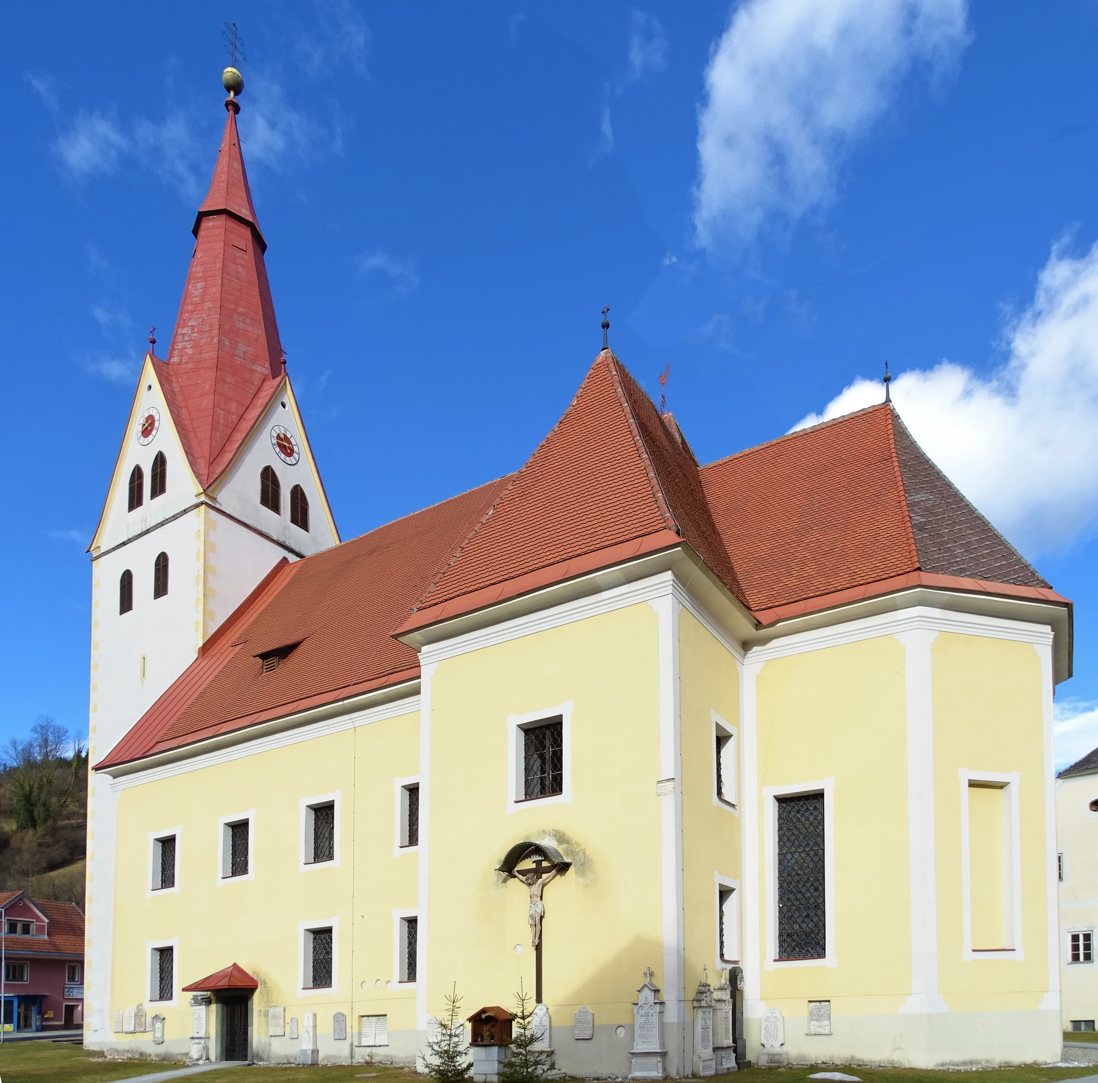 Exterior view of a yellow church with a red spire and tiled roof under a blue sky, featuring a crucifix and gravestones.