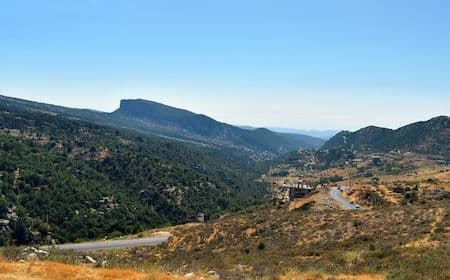 Winding mountain road climbing through a green valley with rocky hills and clear blue sky.