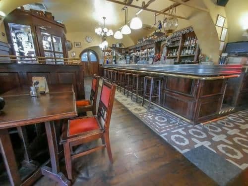 Interior view of a traditional wooden bar featuring a long counter, stools, and dining tables under warm lighting.