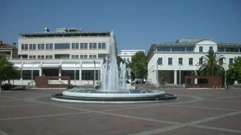 Wide shot of Tuzi's town square with a central fountain, flanked by modern and classical buildings under a clear blue sky.