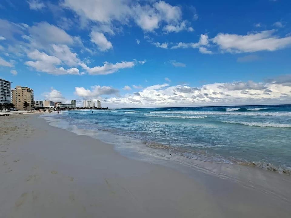 Wide shot of a sandy beach with turquoise waves and a blue sky, featuring distant high-rise buildings along the coastline.