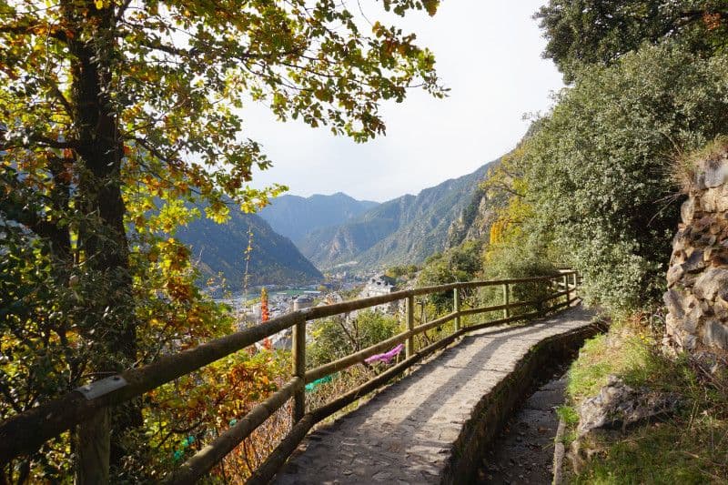 A stone path with wooden railings winds through autumn foliage, overlooking a valley town and mountains.