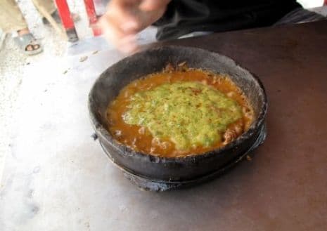 A close-up shot of a sizzling Yemeni street food dish, likely malawach, cooking in a traditional black stone pan with green sauce.