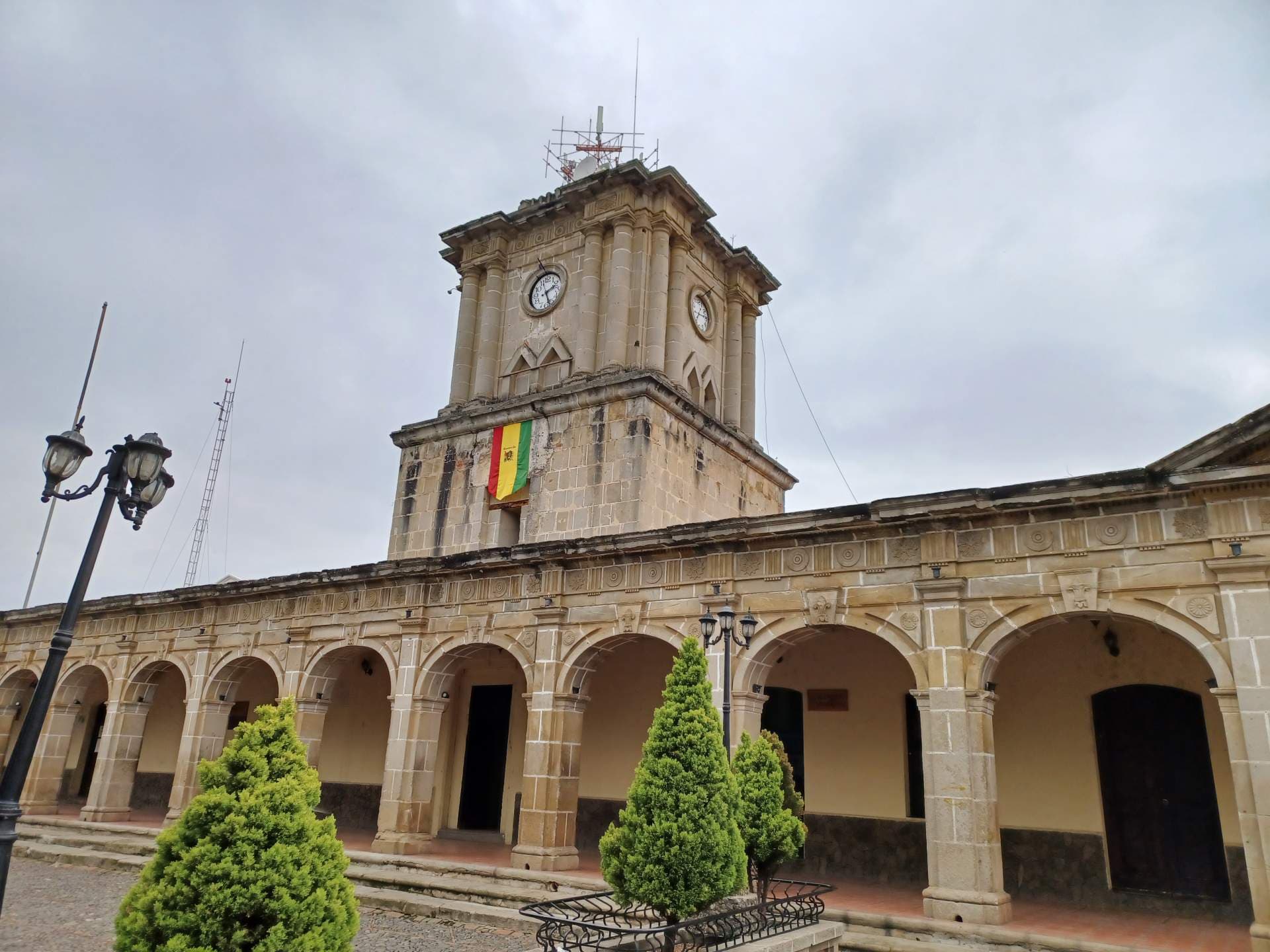 A low-angle shot of a historic stone building featuring a clock tower and arched colonnade, adorned with a Bolivian flag under an overcast sky.