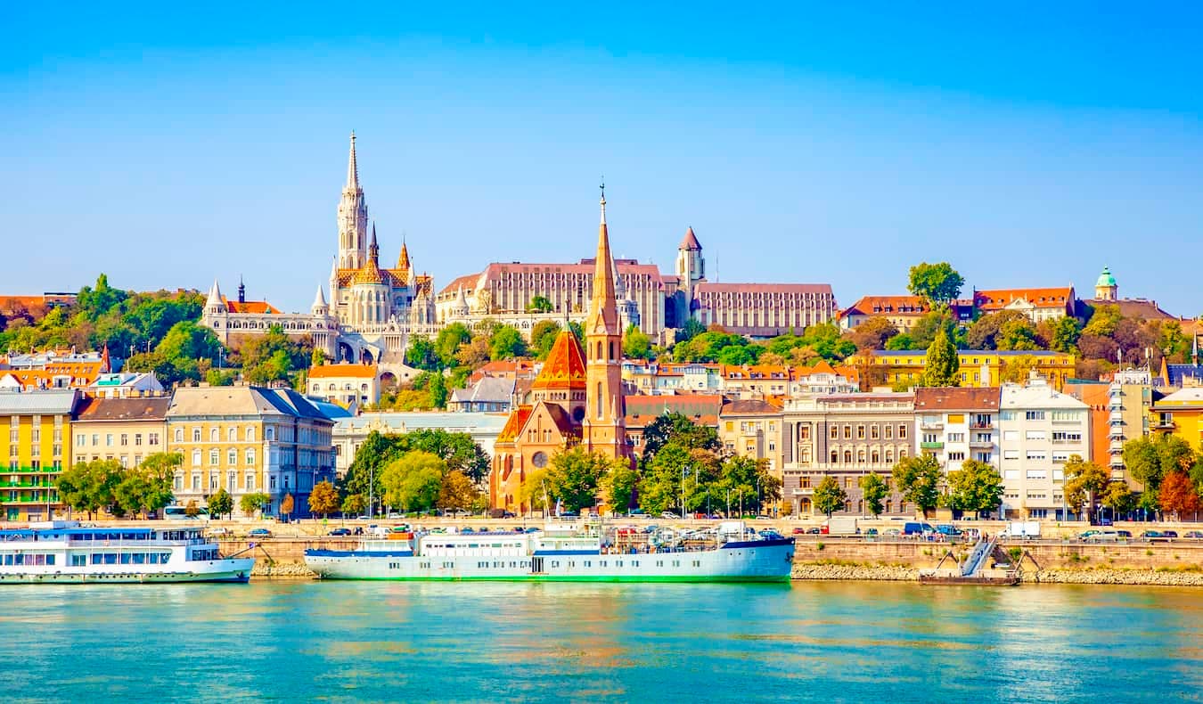 Panoramic view of the Buda Castle District and Matthias Church overlooking the Danube River with cruise ships docked along the waterfront.