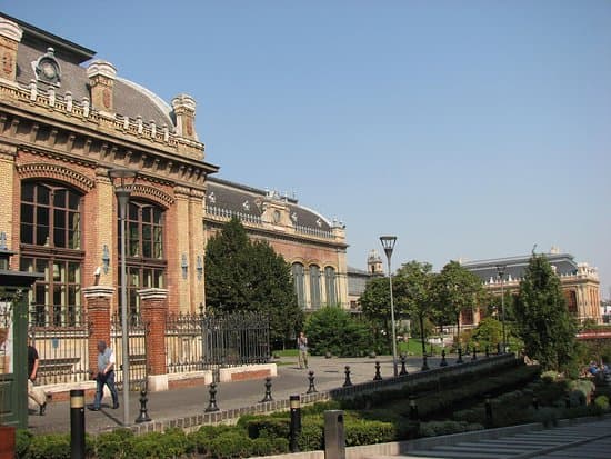 Exterior view of the historic Budapest-Nyugati railway station featuring red brick architecture, arched windows, and a landscaped plaza under a clear blue sky.