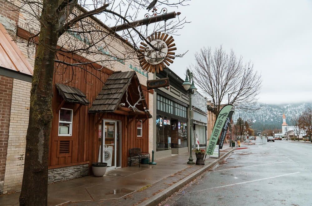 Chewelah main street streetscape with modest storefronts, a hanging sunburst clock, bare trees, and a wet road leading toward distant mountains.