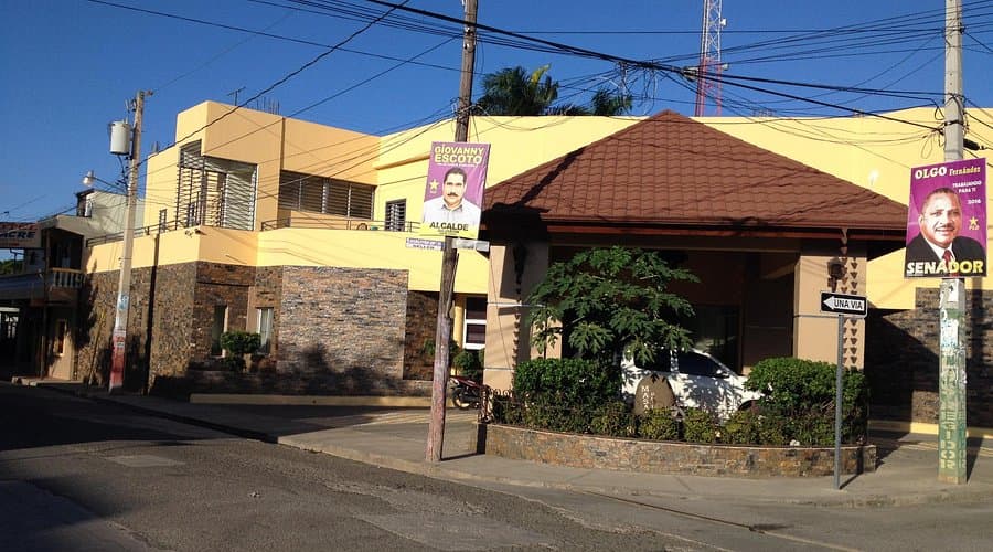 Exterior view of a yellow building with a tiled roof and political campaign posters on utility poles along a street.