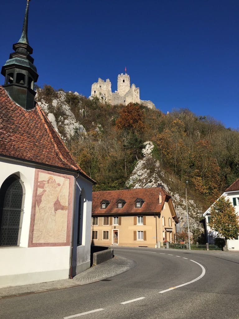 Village church exterior with mural, residential buildings, and a hilltop castle ruin under a clear blue sky near Wolfwil, Canton Solothurn.