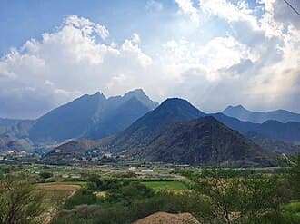 A wide landscape view of the Ta'if valley featuring green agricultural fields in the foreground and rugged mountains under a cloudy sky.