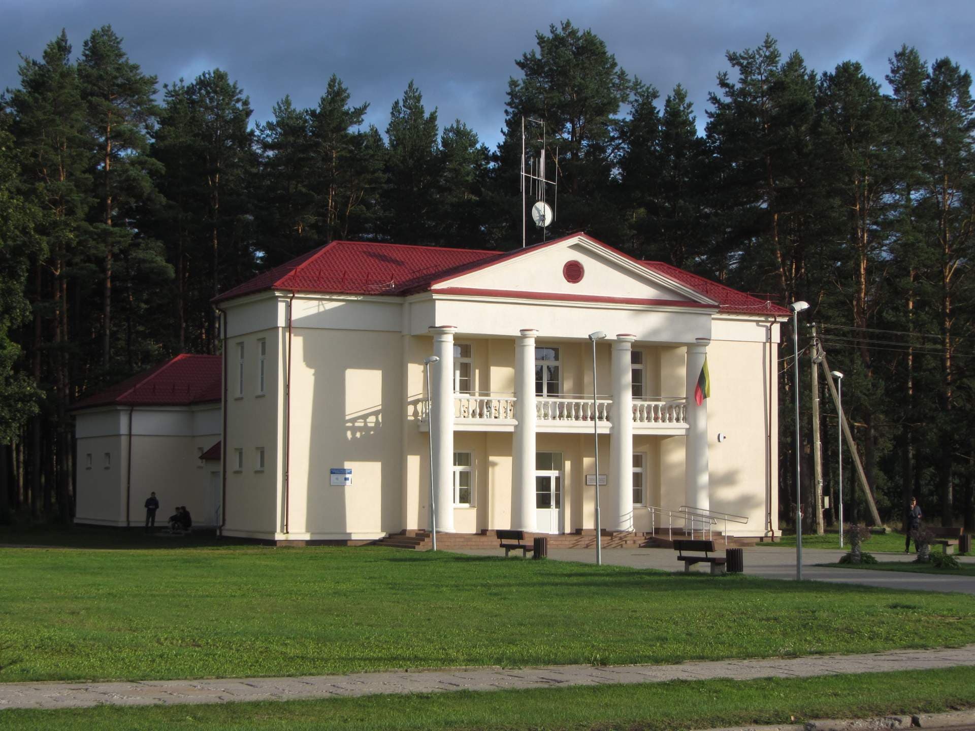 A cream-colored building with a red roof and white columns stands in a grassy area surrounded by tall pine trees under a cloudy sky.