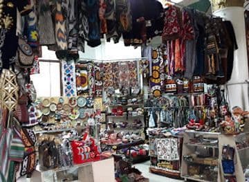 A crowded local market stall displaying hanging textiles, embroidered bags, and shelves of souvenirs and crafts.