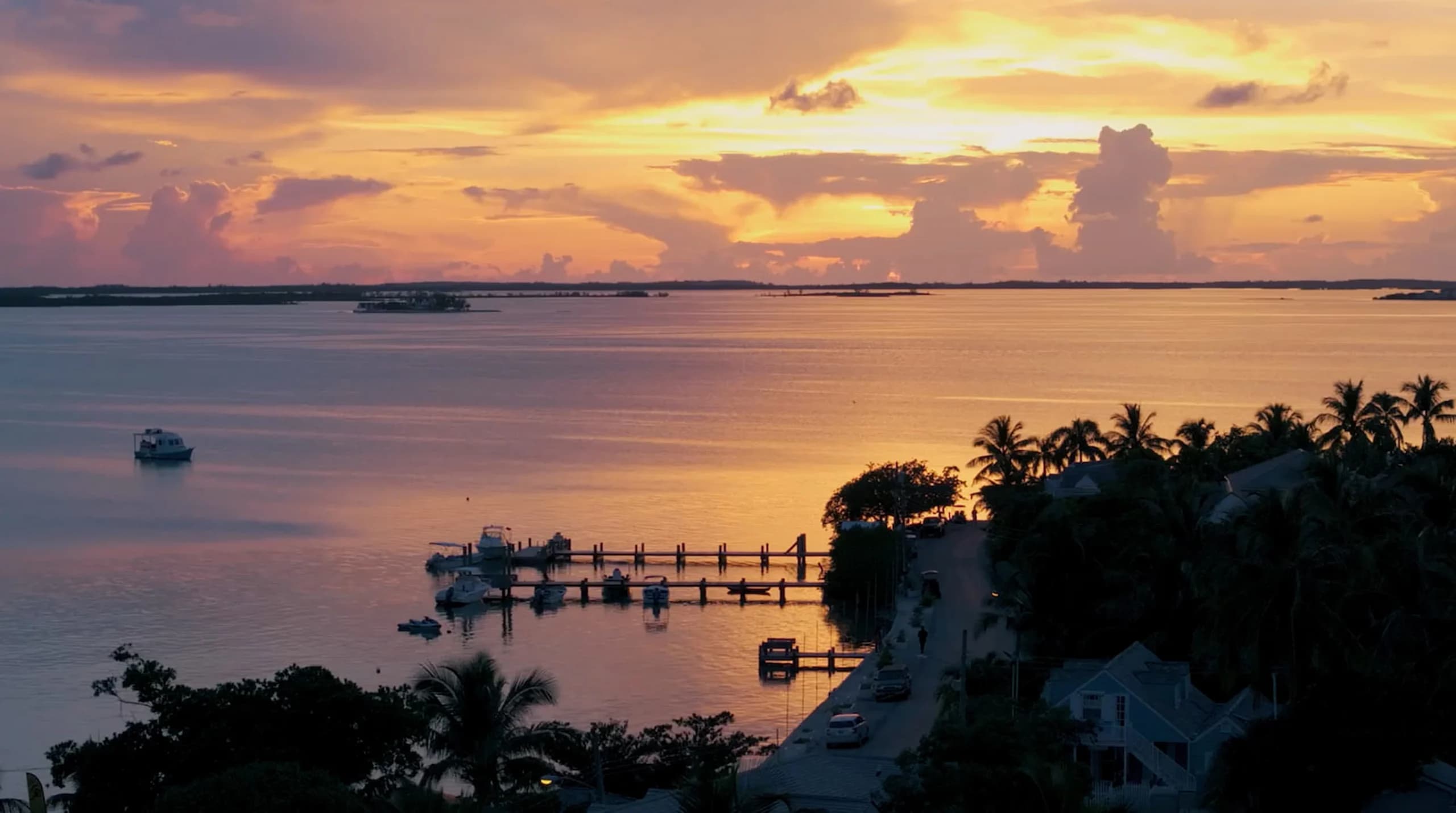 Aerial view of a tropical waterfront at sunset featuring a dock with boats, palm trees, and a coastal road on Abaco Island.