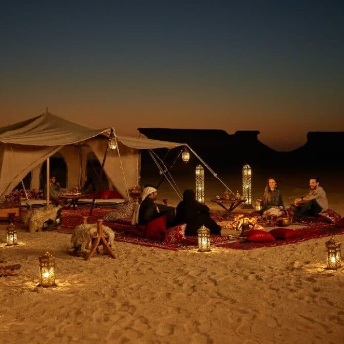 Travelers relax on red carpets around a campfire near a luxury desert tent at dusk, illuminated by lanterns.