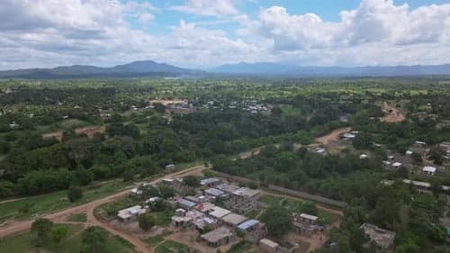 Aerial view of a rural settlement with scattered houses and dirt roads surrounded by lush green vegetation and distant mountains under a cloudy sky.