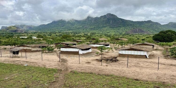 Rural village with simple thatched-roof homes on dirt ground, green hills and cloudy sky in background.