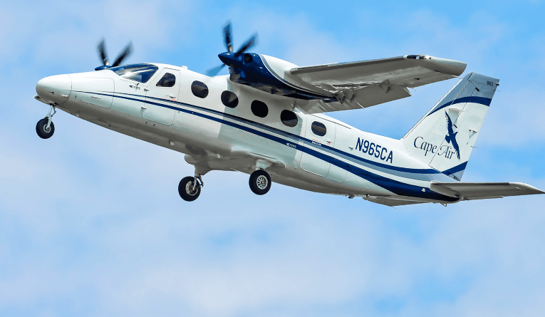 A white Cape Air twin-engine turboprop aircraft with landing gear deployed flies against a blue sky with light clouds.