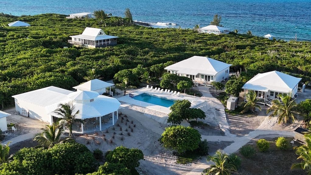 Aerial view of a tropical resort complex with white buildings, a swimming pool, and lush green vegetation near the ocean.