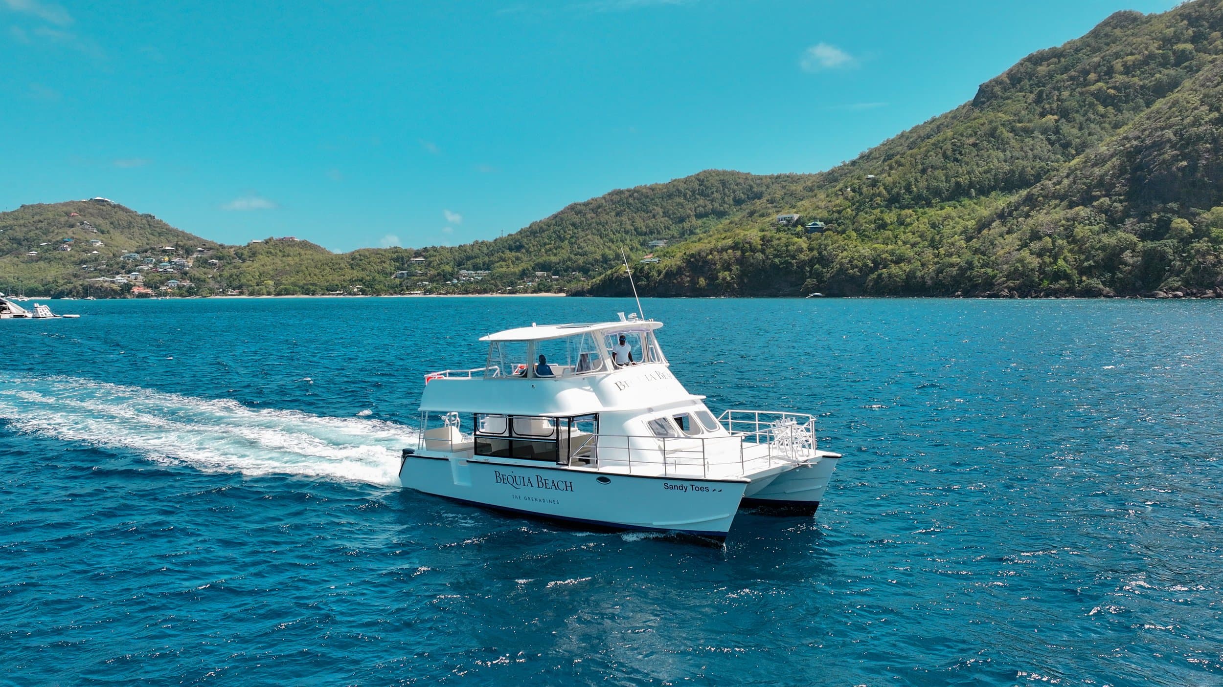 A white catamaran ferry named Bequia Beach cruises through turquoise waters with lush green hills and coastline visible in the background.