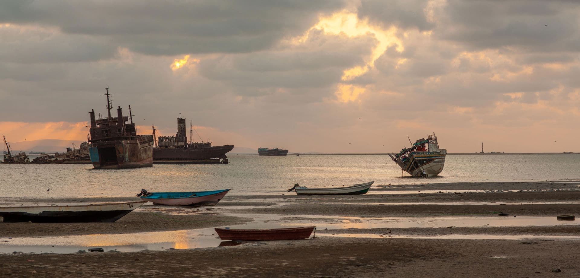 Colorful wooden fishing boats rest on a wet sandy beach at sunset, with rusted shipwrecks and a calm sea in the background.