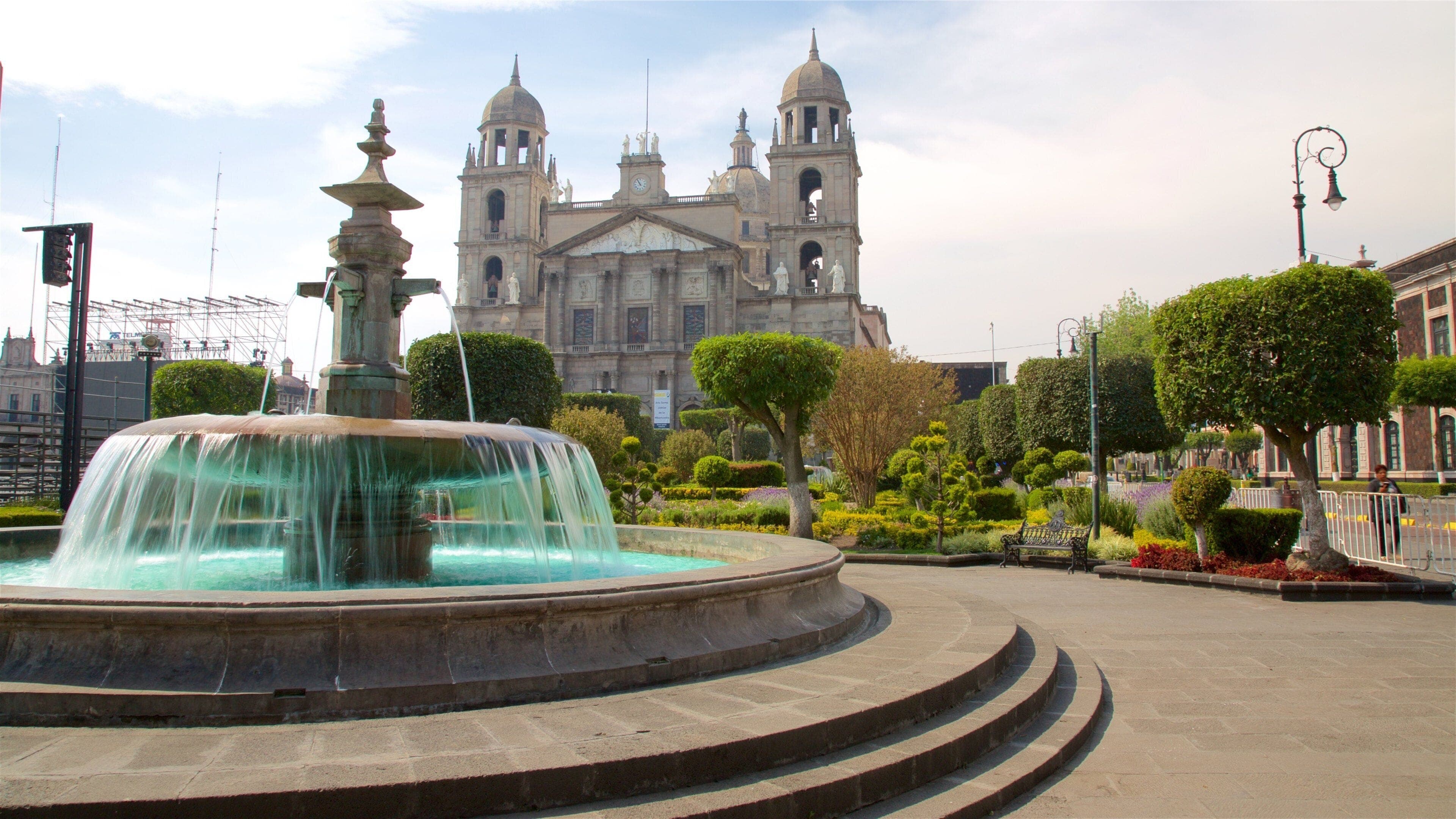 A fountain in a plaza with manicured gardens and a large colonial cathedral in the background under a bright sky.