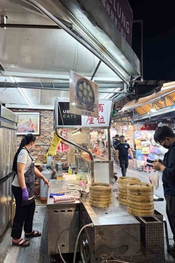 A bustling night market stall in Banqiao features bamboo steamers and a vendor preparing food, capturing the lively street food atmosphere.