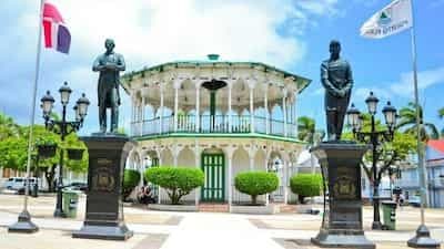 Public plaza in Bisonó featuring a two-story pavilion, statues on pedestals, flags, and ornate street lamps.