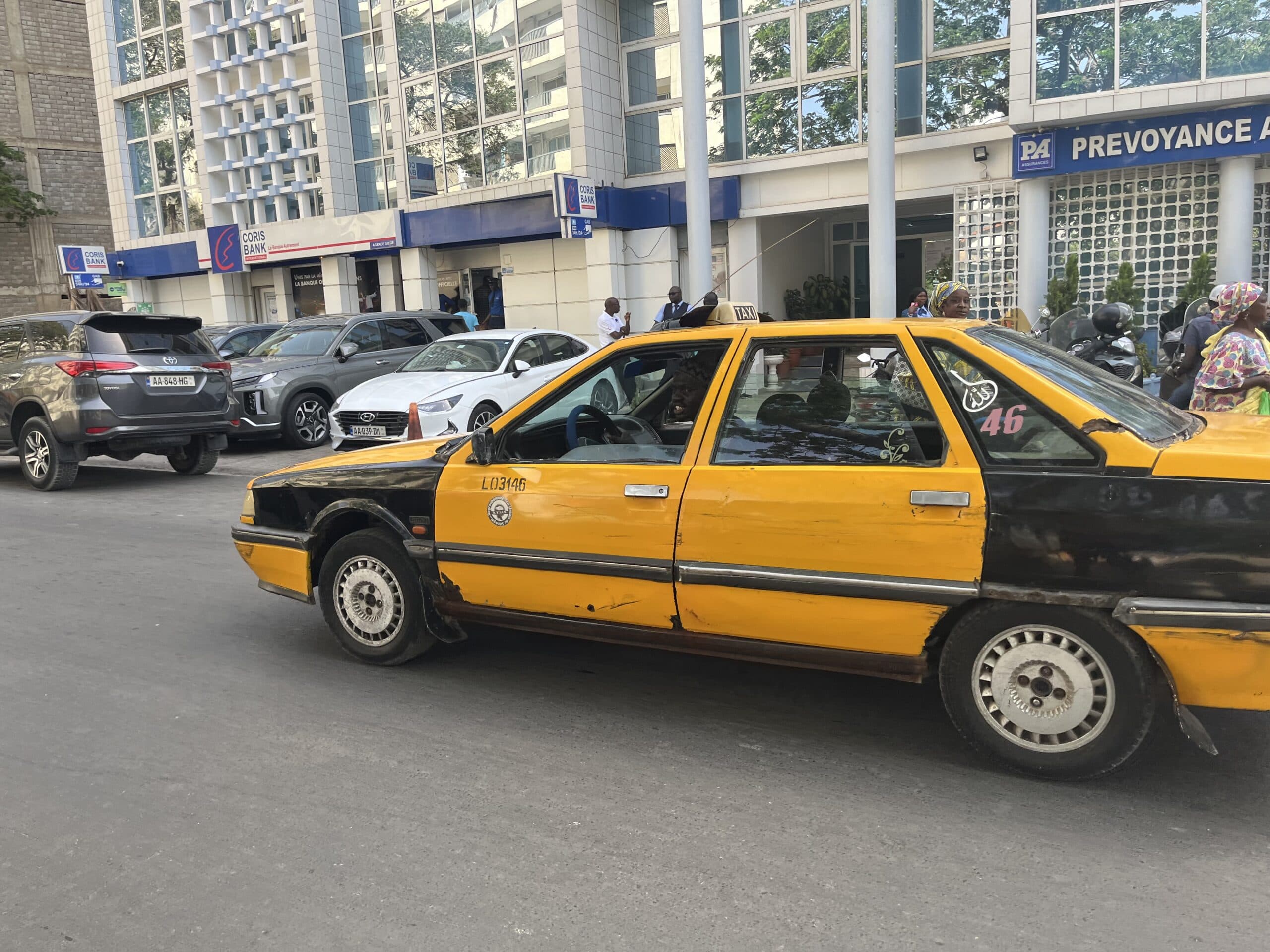 Traffic scene on a coastal road in Dakar, Senegal, with yellow grand taxis and motor scooters navigating the urban environment