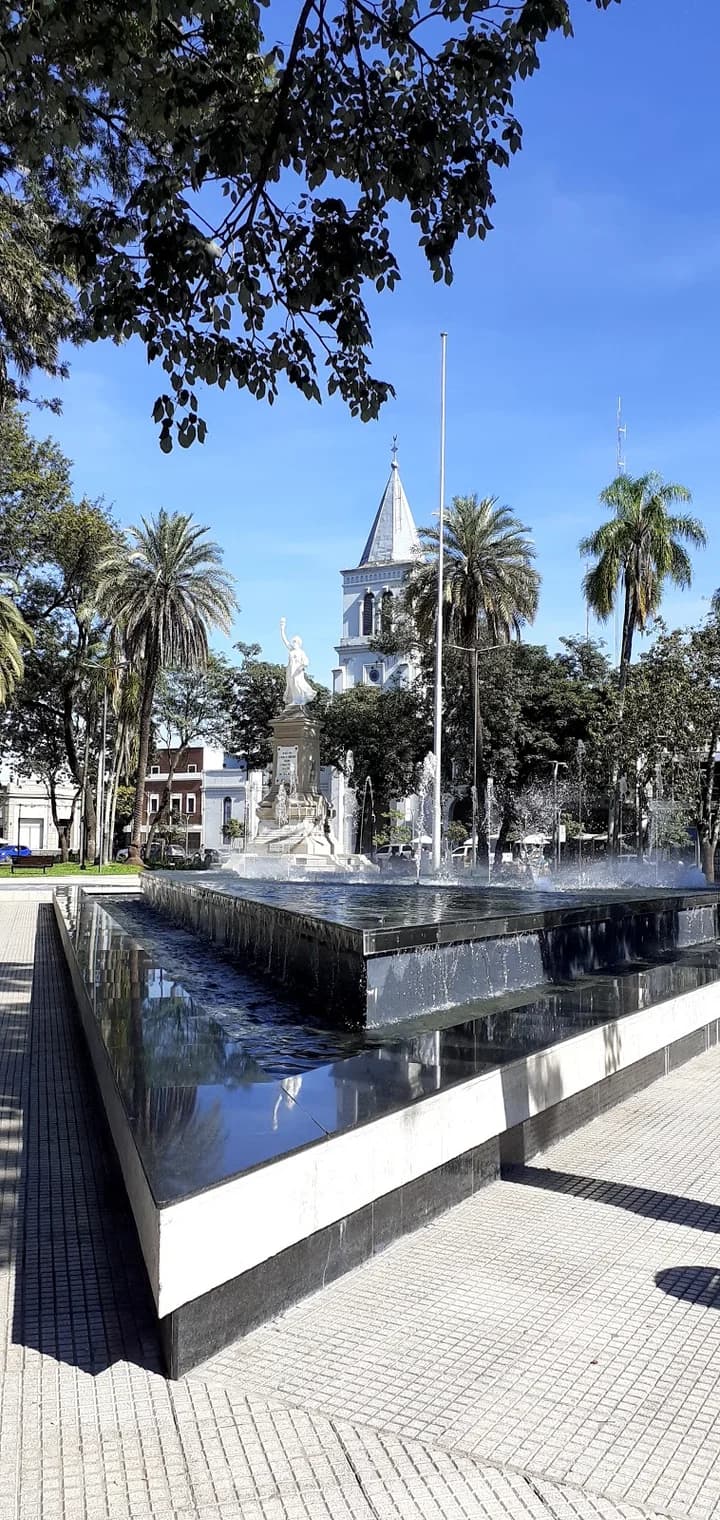 A sunlit plaza in Simoca featuring a modern black fountain, a white statue, palm trees, and a church tower in the background.