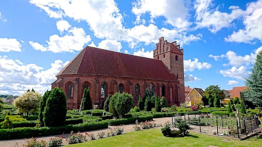 Exterior view of the historic red-brick Præstø Church with its stepped gable tower, set against a blue sky with clouds and surrounded by manicured gardens.