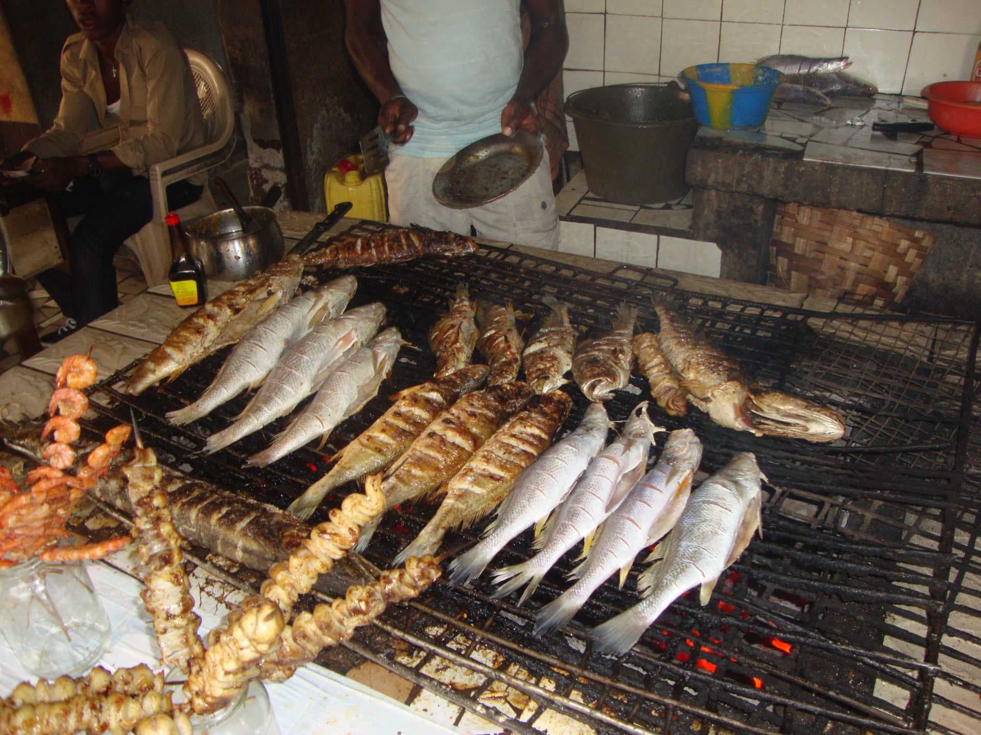 Grilled fish and seafood skewers on a charcoal grill at a beach food stall in Limbe, with vendors in the background