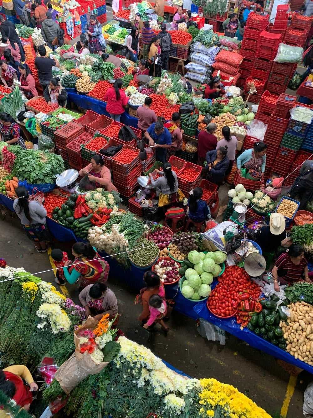 Aerial view of a bustling open-air market with abundant produce, flowers, and goods in crates, surrounded by shoppers and vendors
