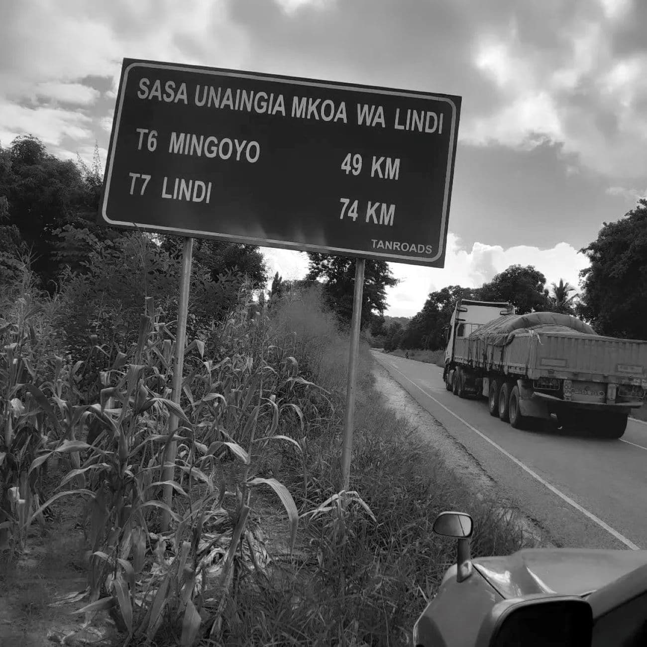 Black and white photo of a road sign entering the Lindi region, listing distances to Mingoyo and Lindi, with a truck driving on the highway.