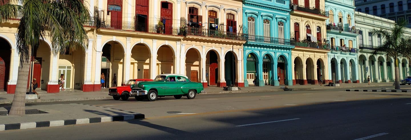 Colorful colonial buildings with arched walkways line a quiet street in San Antonio de los Baños, featuring vintage cars and palm trees.