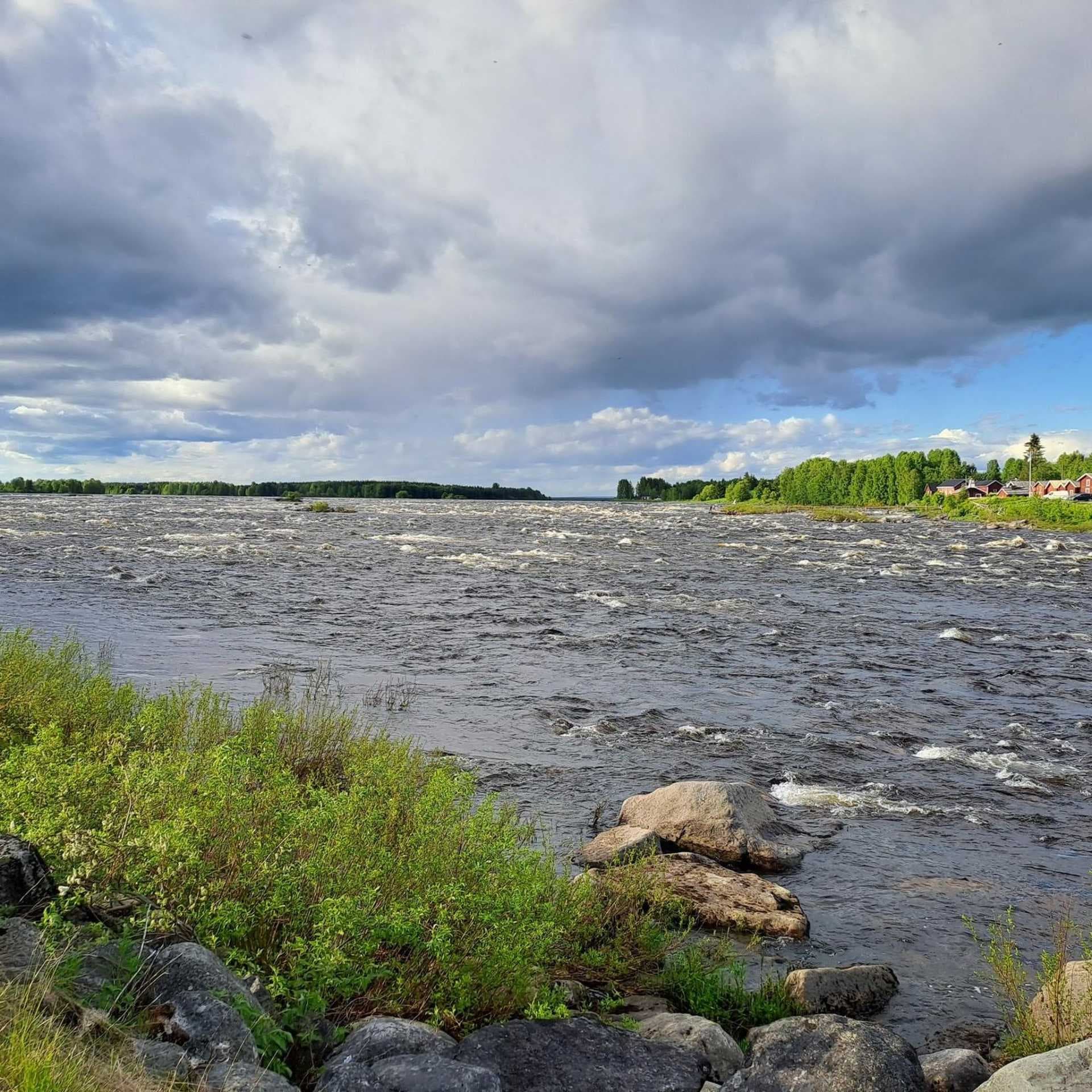 Wide river landscape with turbulent water, rocky foreground, and distant red wooden houses under a cloudy sky.